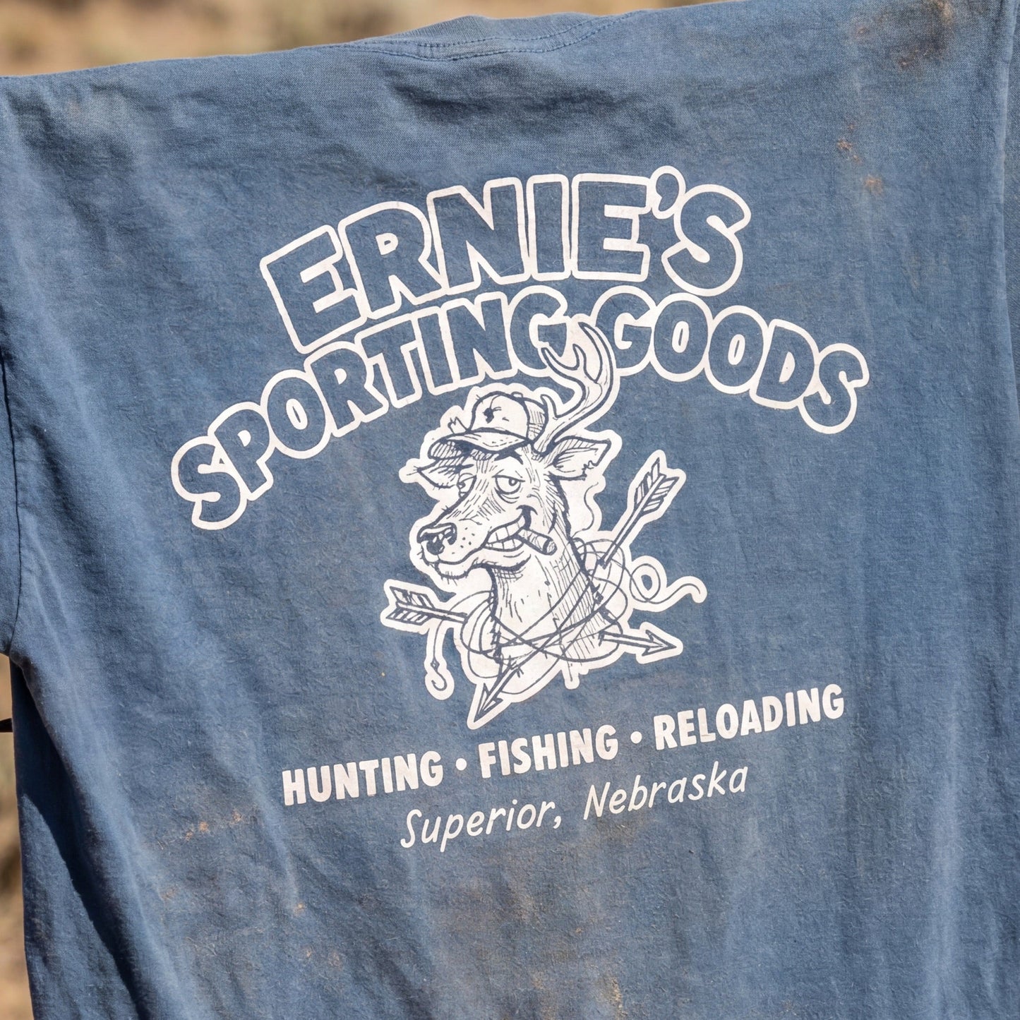 Blue t-shirt with 'Ernie's Sporting Goods' print hanging on a barbed wire fence in a desert setting.