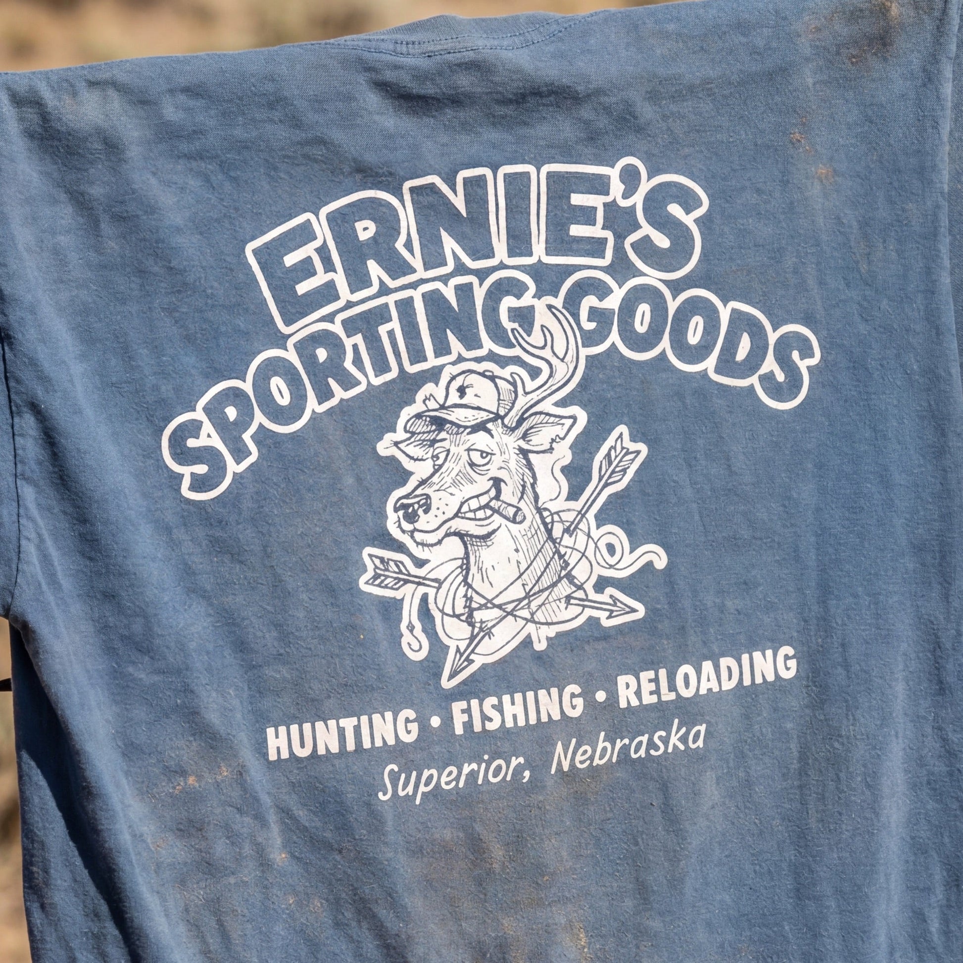 Blue t-shirt with 'Ernie's Sporting Goods' print hanging on a barbed wire fence in a desert setting.