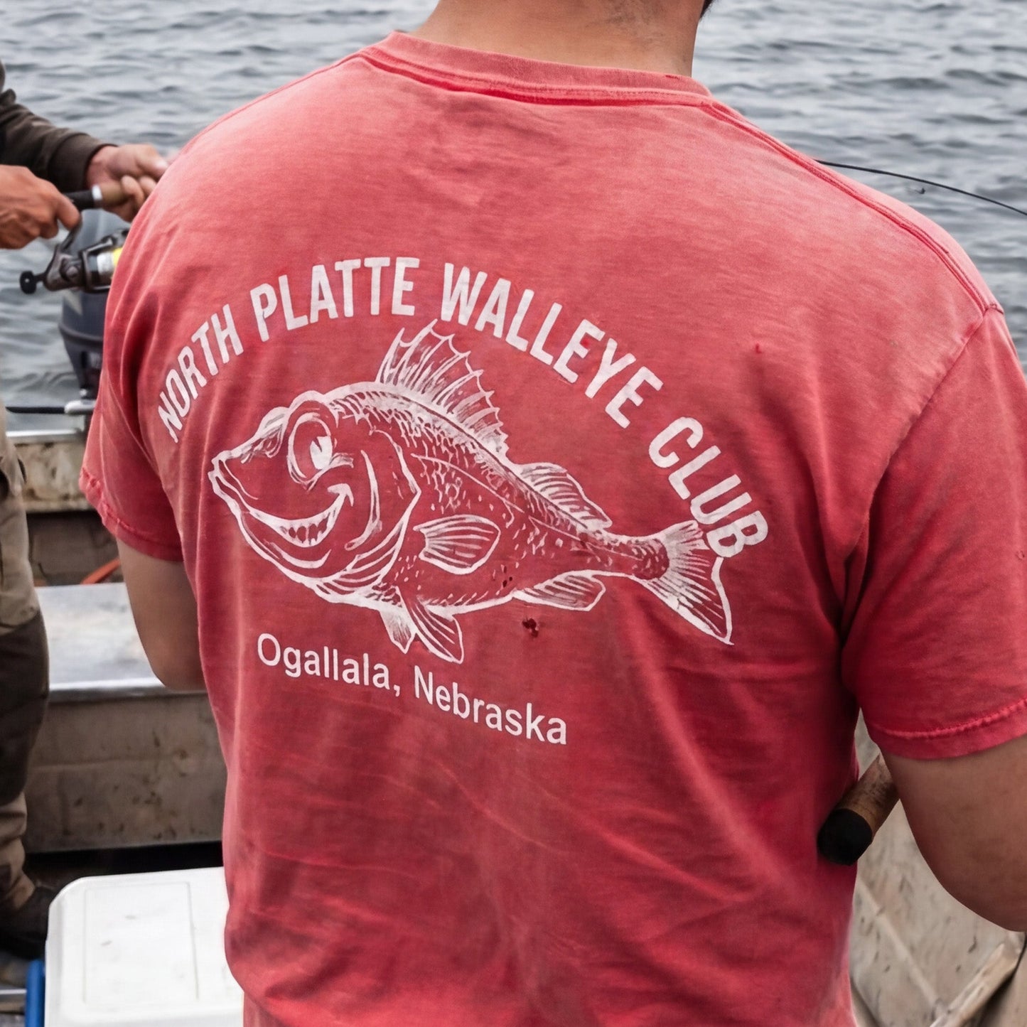 Person wearing a red 'North Platte Walleye Club' t-shirt on a boat by a body of water.