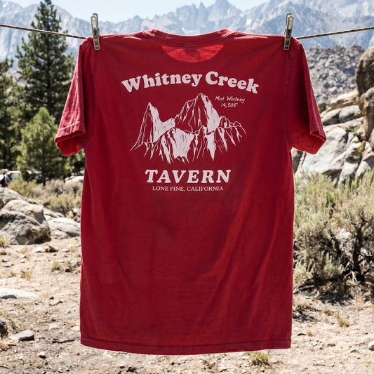 Red t-shirt with 'Whitney Creek Tavern' design hanging on a clothesline with mountains in the background.